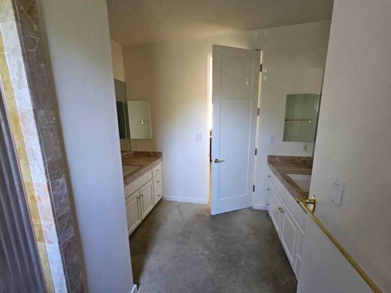 A modern bathroom featuring two white vanities with brown marble-look countertops, a concrete floor, and freshly painted white walls. A white paneled door is slightly open, revealing a bright light beyond. Brass fixtures are visible on the vanities and a towel bar.