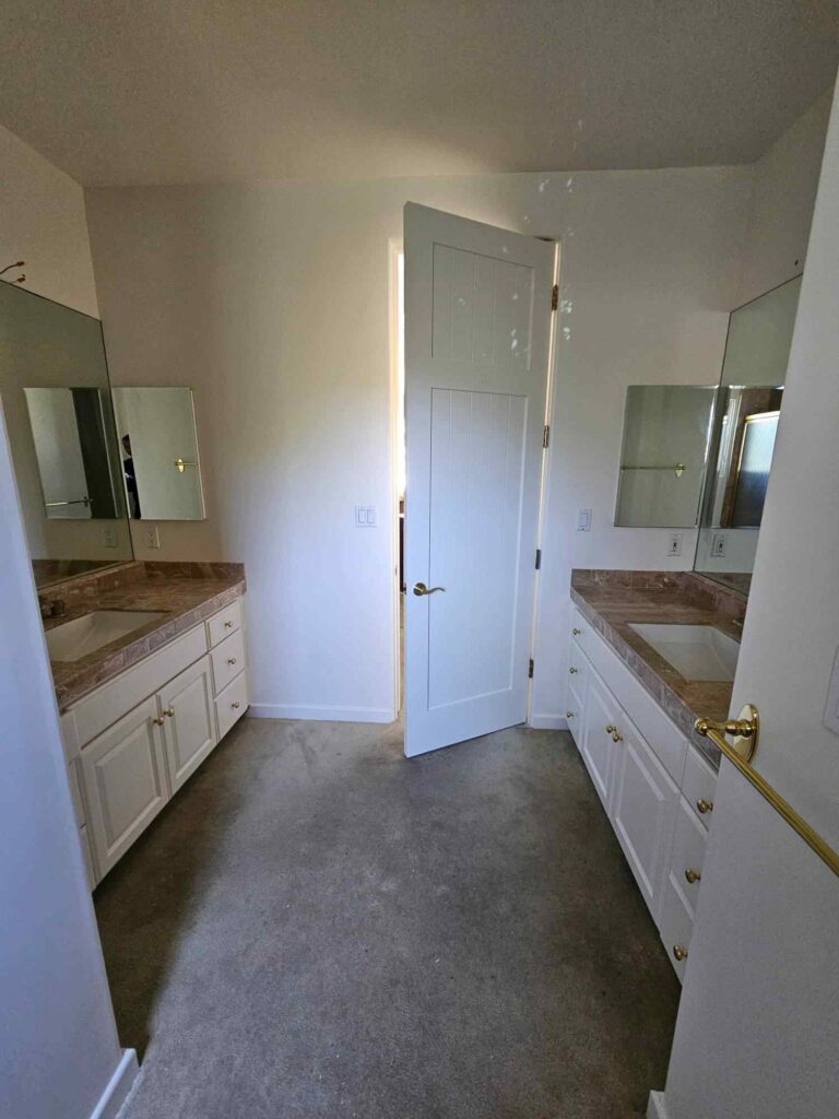 A bright bathroom featuring two white vanities with marble countertops and white cabinets, flanking a partially open white door. The walls are painted a clean white, and the floor is carpeted.