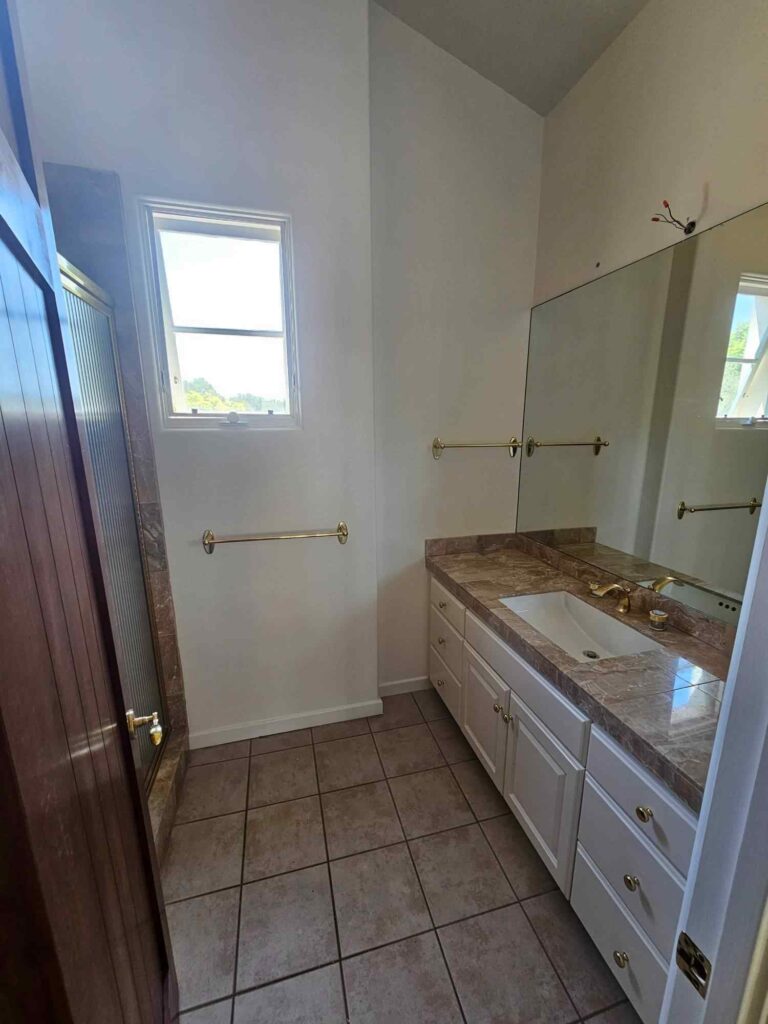 A spacious bathroom featuring a white vanity with a brown marble countertop and a white sink, gold fixtures, and a large wall mirror. To the left, a shower with a glass door and brown tile accents is visible next to a wooden door. A window above a gold towel bar lets in natural light, illuminating the light-colored walls and brown tiled floor. Exposed wires are visible above the mirror, indicating a potential renovation project.