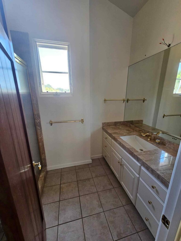 A bathroom featuring a white vanity with a brown marble countertop, a white sink, and gold fixtures. A large mirror hangs above the vanity, reflecting a window that lets in natural light. The floor is covered with brown tiles, and a wooden shower door is visible on the left.