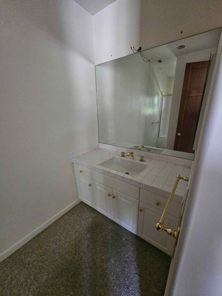 A small bathroom with a white vanity, tiled countertop, gold faucet, and a large mirror. The plain white walls show exposed wiring above the mirror, indicating a need for painting or renovation. The floor is carpeted.