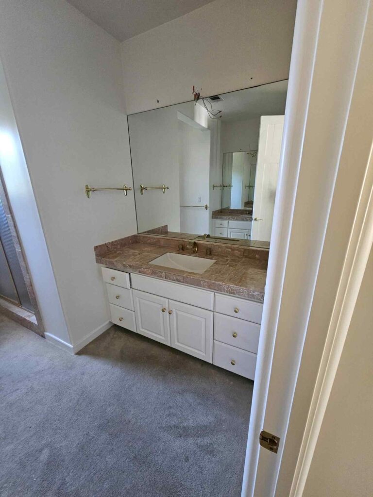 A bathroom featuring a white vanity cabinet with a light brown marble countertop, a sink, and a large mirror. White walls are visible, along with brass towel bars and cabinet knobs. Exposed wires above the mirror indicate a painting or renovation project in progress.