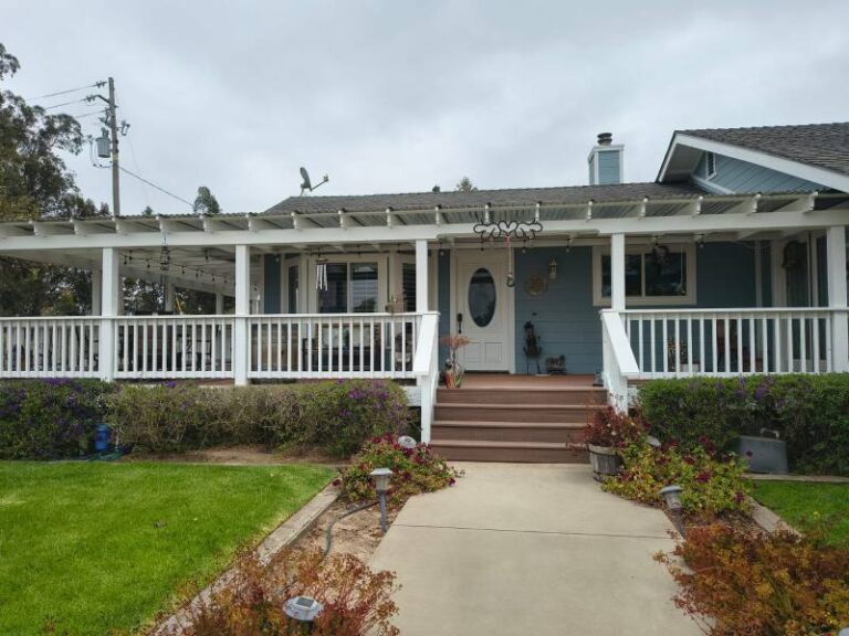 Exterior view of a light blue house with a wide white porch, white railings, and brown steps leading to a white front door. The house is surrounded by green grass and landscaping under a cloudy sky.