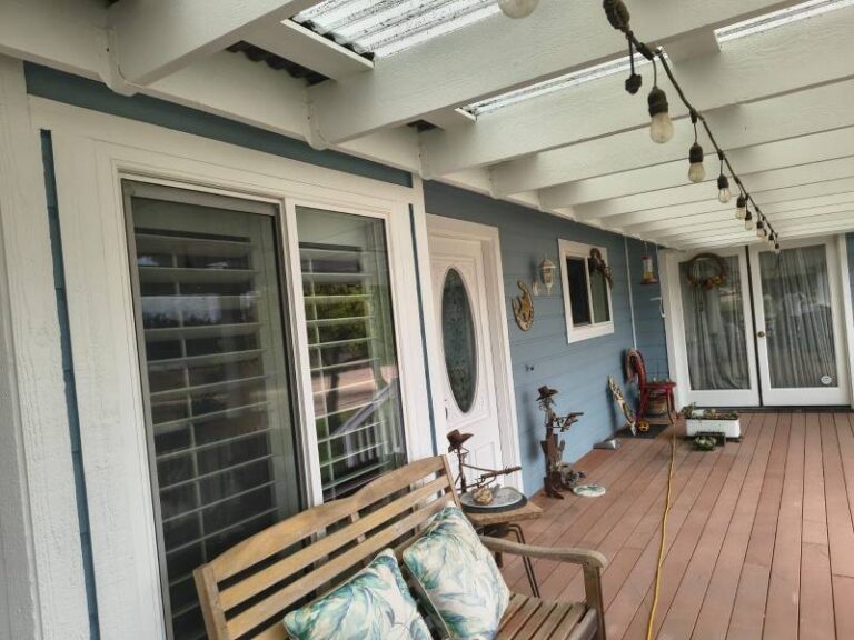 A welcoming house porch featuring light blue siding and crisp white trim around windows and doors, showcasing a fresh paint job. A wooden deck and bench with decorative pillows are visible, along with string lights overhead.