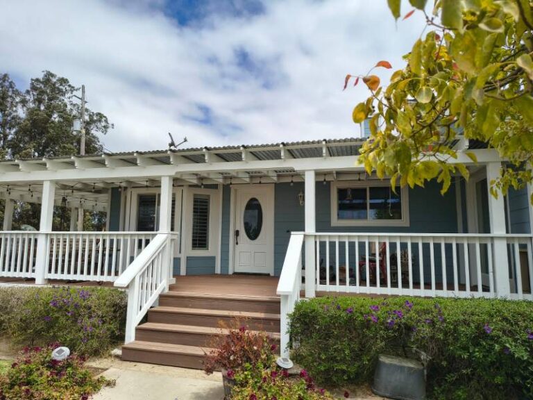 A house with a freshly painted blue exterior, white porch railing, and brown steps, surrounded by green shrubs and flowering plants, showcasing quality exterior paintwork.