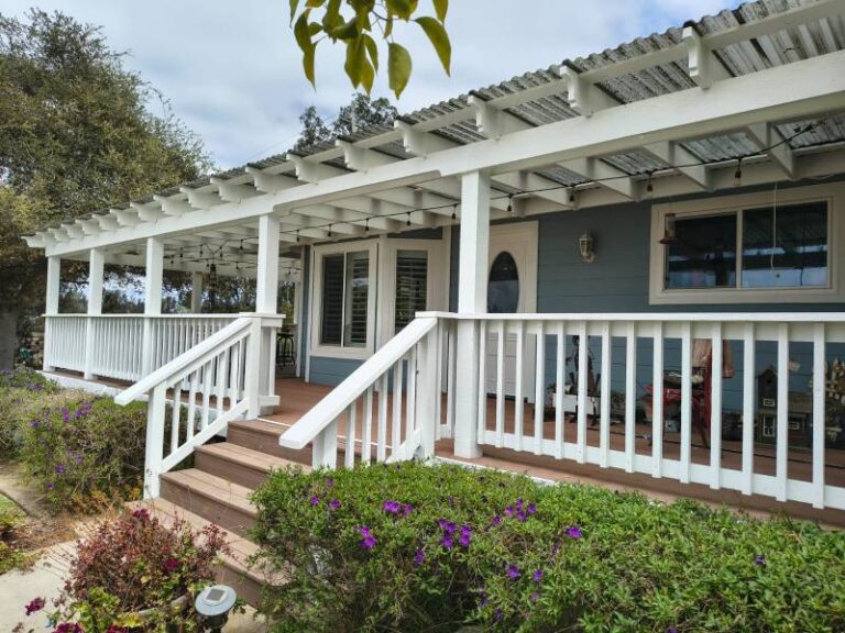 Exterior view of a house with light blue siding, a freshly painted white wooden porch with railing and steps, and a white pergola roof with string lights, surrounded by green bushes with purple flowers.