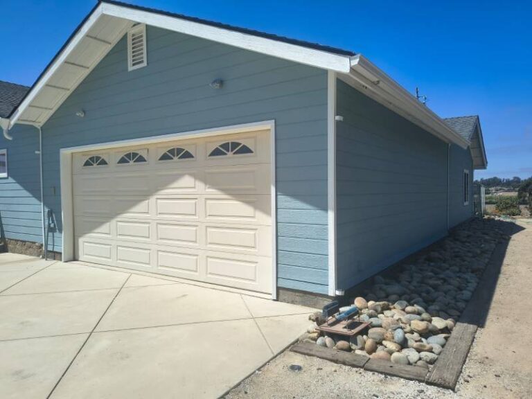 Exterior view of a house with light blue siding, white trim, and a beige garage door with arched windows. A concrete driveway leads to the garage, and a decorative rock bed runs along the side of the house under a clear blue sky.