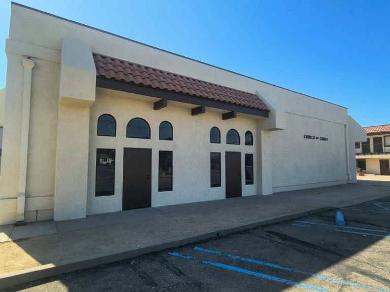 Exterior view of a Church of Christ building with light beige stucco walls, dark brown doors, arched windows, and a Spanish tile roof accent under a clear blue sky. A parking lot with blue markings is in the foreground.