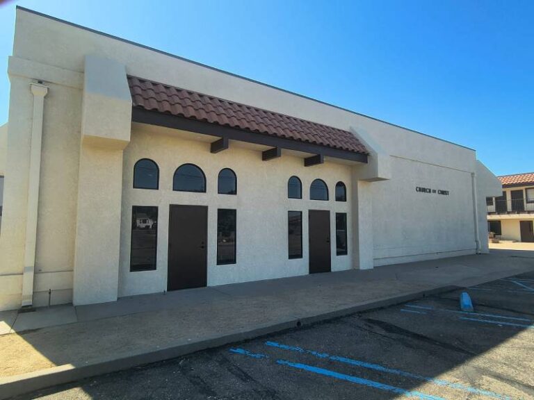 Exterior view of a Church of Christ building with light beige stucco walls, a brown tiled roof, and dark brown doors with arched windows, under a clear blue sky. An asphalt parking lot with blue markings is in the foreground.