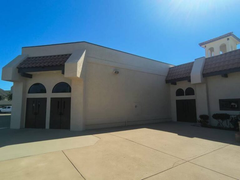 Exterior view of a light-colored commercial building with stucco walls, dark brown tile roofs, and dark double doors with arched windows, under a bright blue sky. A concrete patio is in the foreground.