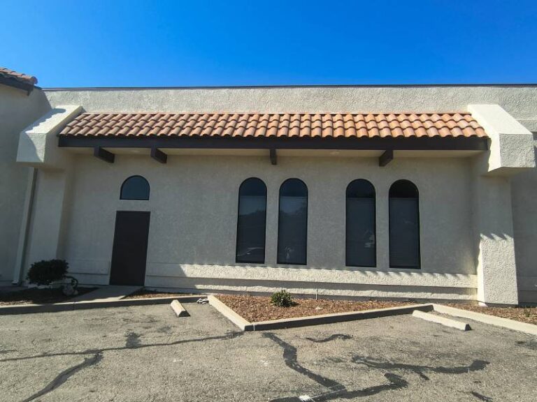 Exterior view of a commercial building with light stucco walls, a brown tile roof overhang, and arched windows. A small parking lot with cracks is in the foreground.