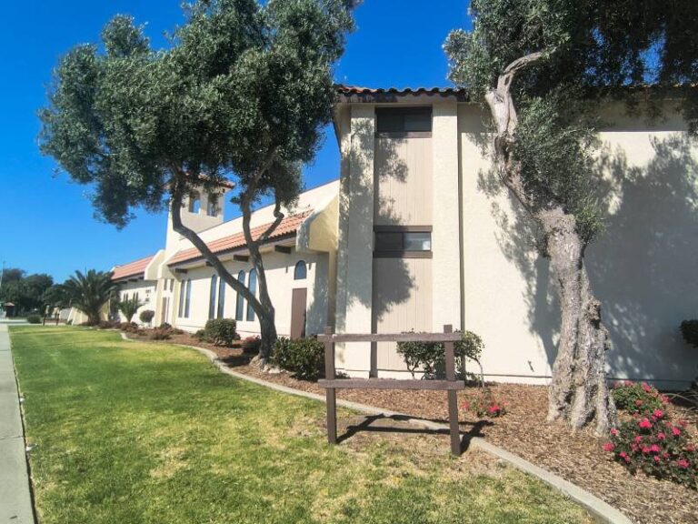 Exterior view of a newly painted commercial building featuring clean off-white stucco walls, neutral trim, and terracotta roof accents surrounded by green landscaping.