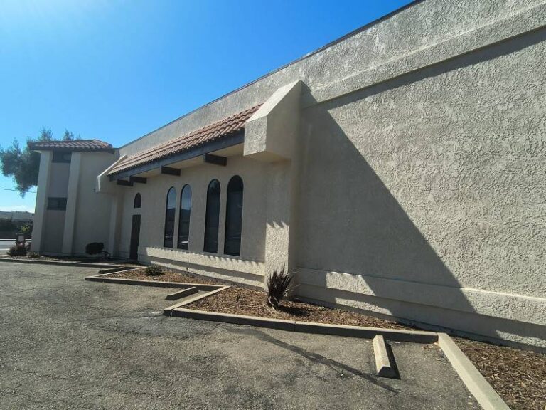 Exterior view of a commercial building featuring a clean beige stucco finish and arched windows under a bright blue sky.