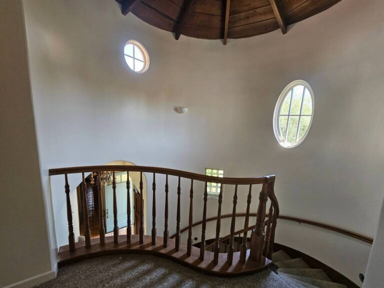 A view looking down a curved wooden staircase with a carpeted tread and a matching wooden balustrade. The staircase is set against a light-colored, round wall with an oval window and a smaller round window. A wooden, domed ceiling is visible above, and an arched doorway in the background reveals a light blue door.