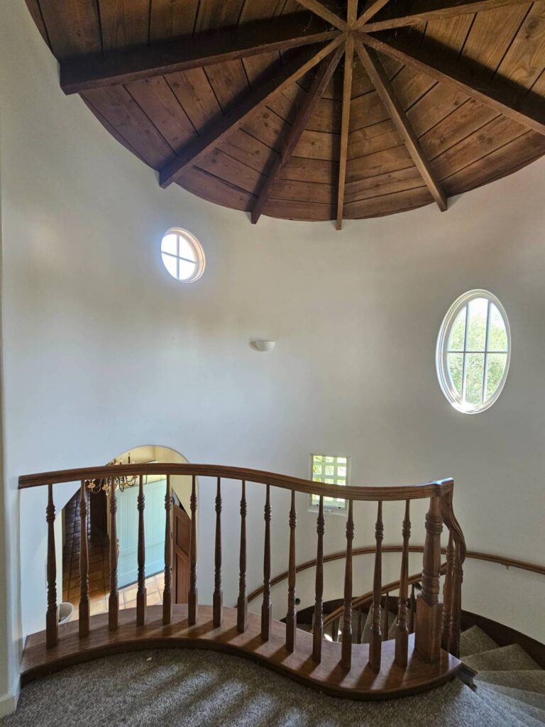View from above a curved staircase with a wooden railing and carpeted steps, leading down. The white, curved walls feature a small round window and a larger oval window, letting in natural light. Above, a rustic wooden ceiling with radial spokes completes the unique interior design.