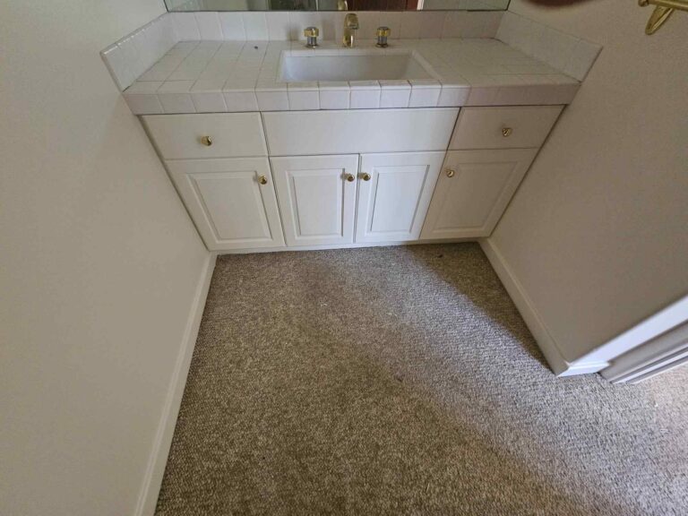 A dated bathroom vanity featuring a white tiled countertop with an integrated sink, gold-colored faucet, and white cabinets with gold knobs. The floor is covered with light brown carpet, and the walls are a light neutral color.