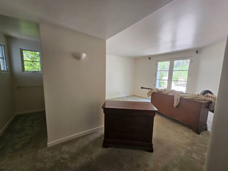 An empty bedroom featuring freshly painted white walls and a light green carpet. A dark wooden dresser stands in the foreground, while a wooden bed frame draped with a striped cloth is positioned near French doors that open to an outdoor area with trees. A small alcove with a window is visible on the left.
