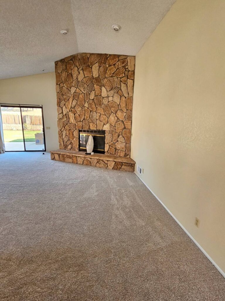 Empty living room with a large corner stone fireplace, light beige painted walls, and new beige carpet. A sliding glass door leads to a green backyard.