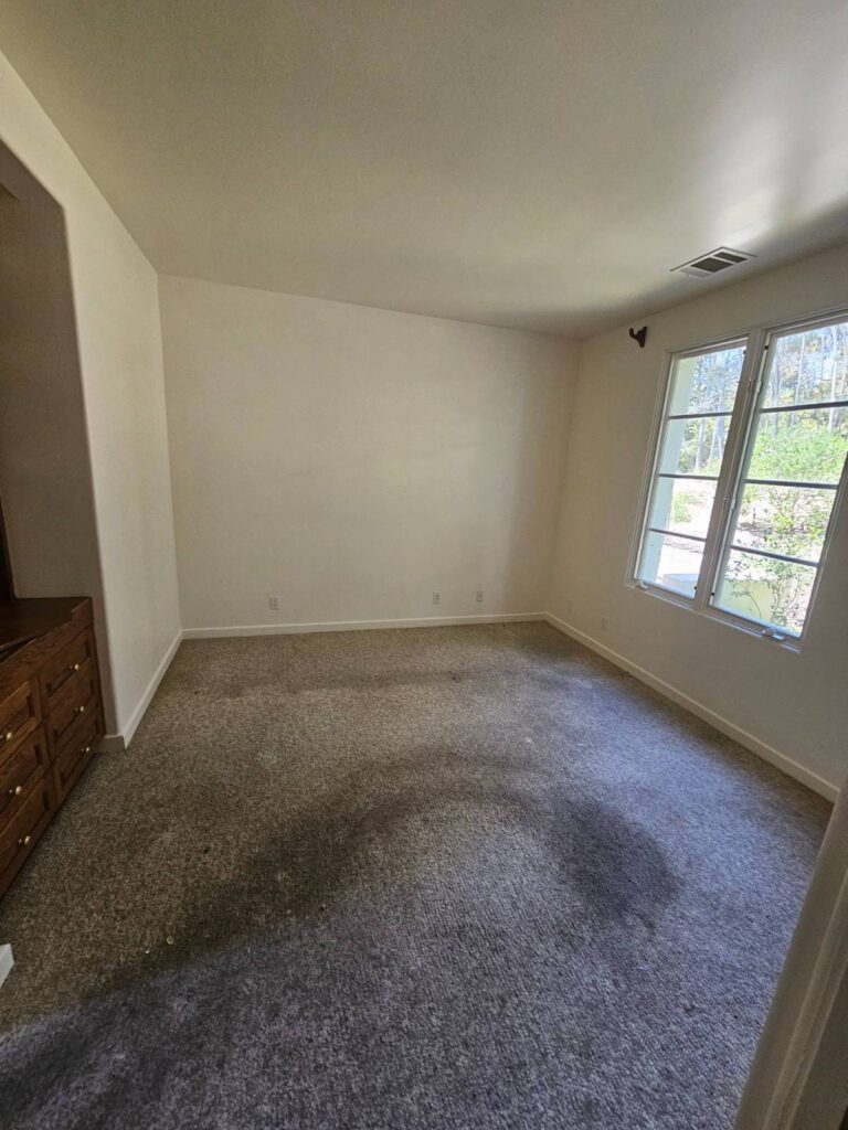 An empty residential room with light beige walls, a worn grey carpet, a large window looking out to trees, and a wooden dresser on the left. The room appears ready for painting.