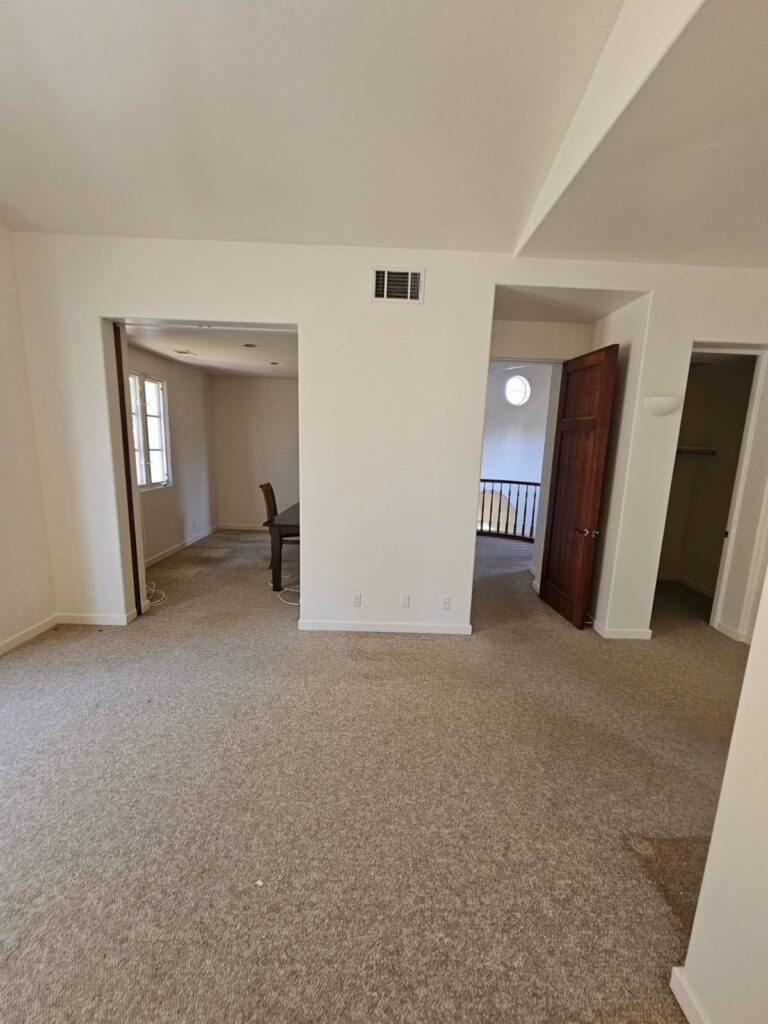 An empty room with light beige carpet and freshly painted white walls, featuring multiple doorways leading to other areas including a dining room and a staircase. The room is ready for new decor or further painting.