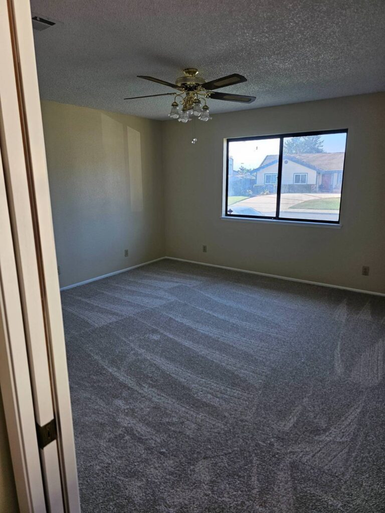An empty residential room featuring light beige painted walls, a textured popcorn ceiling, a brass ceiling fan with glass light shades, and new gray patterned carpet. A large window offers a view of a street and neighboring houses. The room is clean and ready for painting or further decoration.