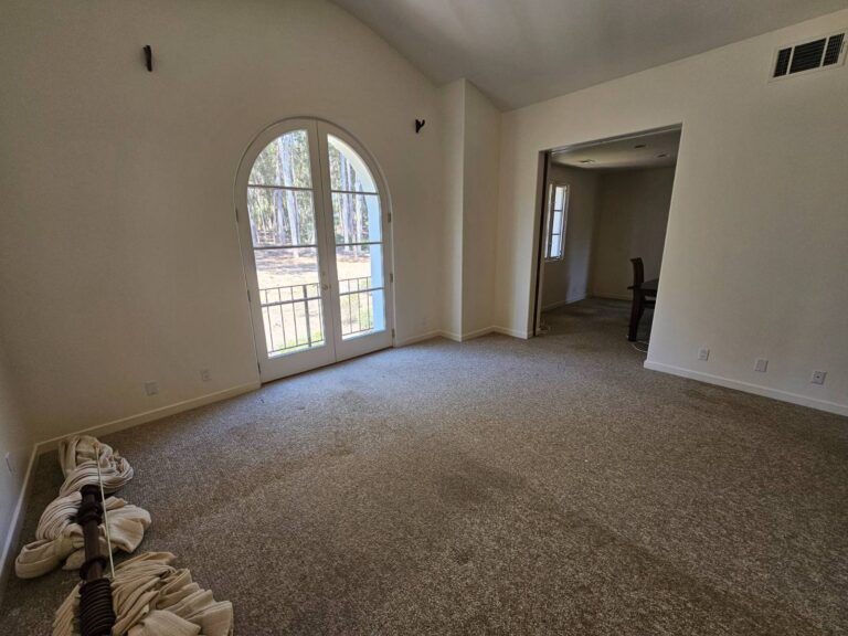 Empty residential room with light walls, vaulted ceiling, and an arched double door leading to an outdoor view of trees. Light brown carpet covers the floor, and an opening to another room is visible on the right. Painting supplies are in the bottom left.