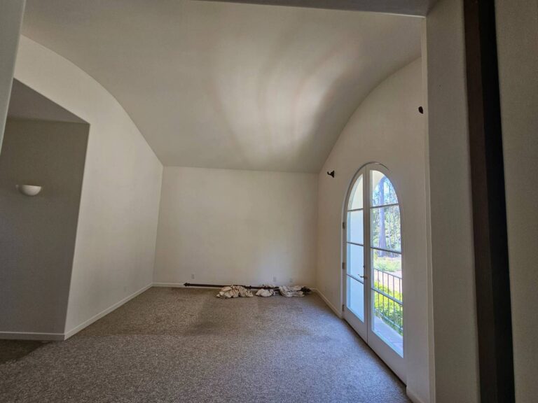 An empty room with freshly painted white walls, a vaulted ceiling, and light gray carpet. An arched glass double door on the right opens to an outdoor view of trees and greenery. A white wall sconce is visible on the left wall, and some rolled-up materials are on the floor against the far wall, indicating a space ready for painting or renovation.