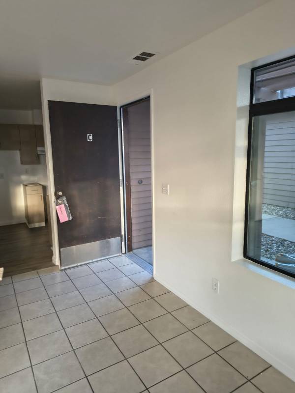 An interior entryway featuring freshly painted white walls, a light-colored tile floor, and a dark brown door with a 'C' on it. A window on the right looks out to an outdoor area, and a kitchen with light wood cabinets is visible in the background.
