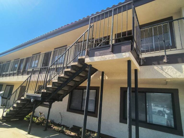 Exterior of a two-story apartment building featuring freshly painted beige stucco walls with dark brown window trim and metal railings.