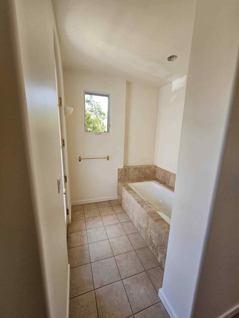 A freshly painted bathroom with light beige walls, a built-in bathtub surrounded by brown marble-look tiles, a small window, a gold towel bar, and light brown square floor tiles.