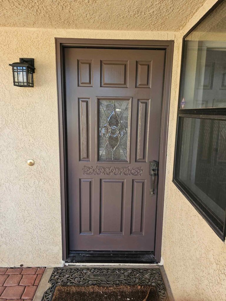 A freshly painted brown front door with decorative glass and a black handle, set in a light beige stucco wall. A black lantern light fixture is on the left, and a window is on the right. A welcome mat rests on brick pavers in front of the door.