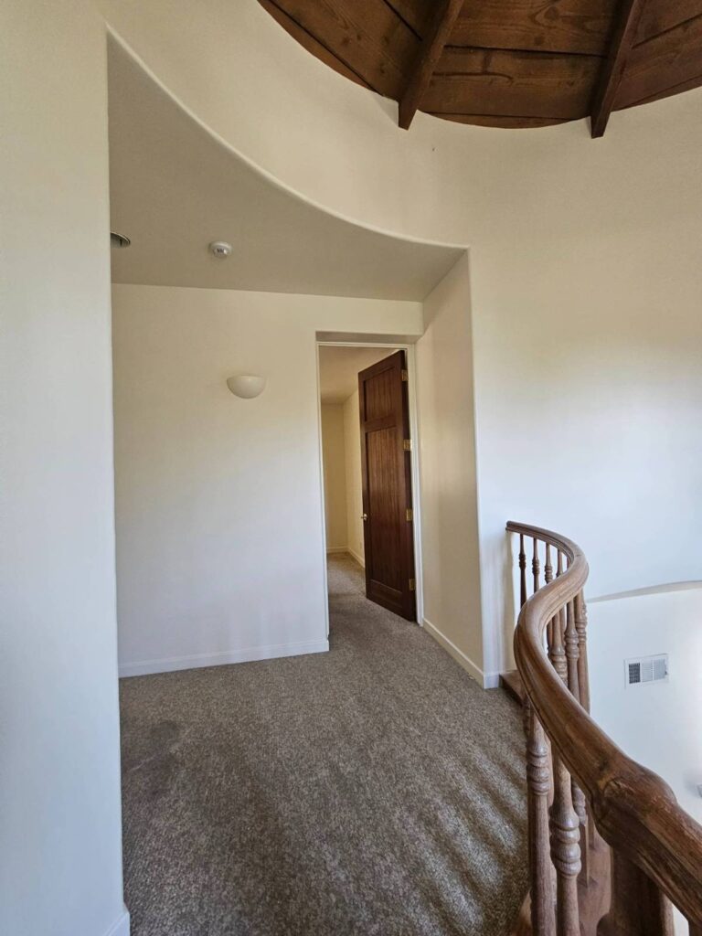 Freshly painted interior hallway with clean white walls, light brown carpet, a dark wood door slightly ajar, a curved wooden railing, and a rustic wooden ceiling.