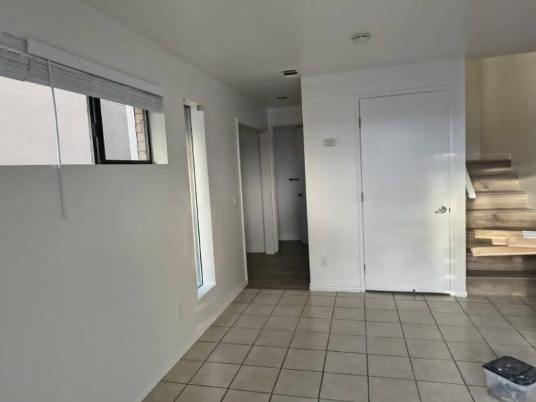 An empty interior room with freshly painted white walls, a light-colored tiled floor, and two windows with blinds on the left. A white door is visible on the right, next to a wooden staircase. The room appears clean and ready for occupancy or further decoration.