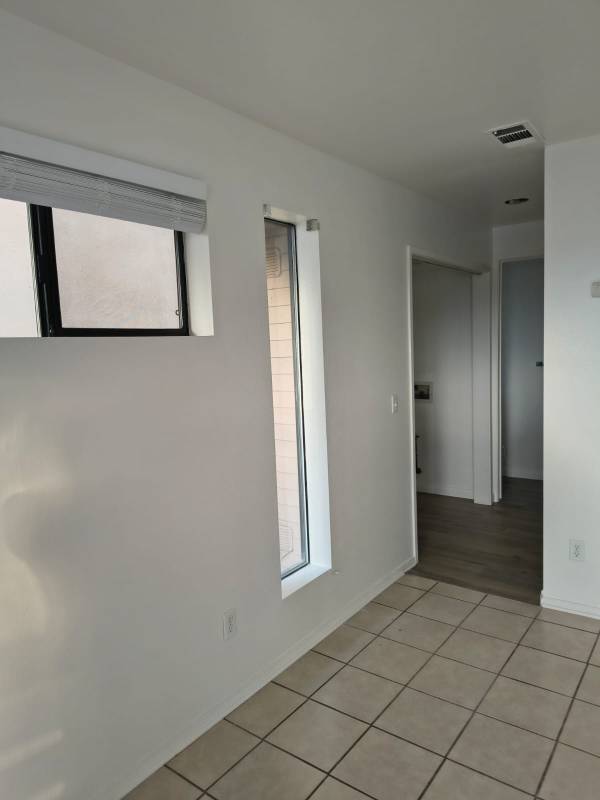 An interior room with freshly painted white walls, featuring a horizontal window with blinds and a vertical window, light beige tile flooring, and a doorway leading to a room with wood-look flooring.