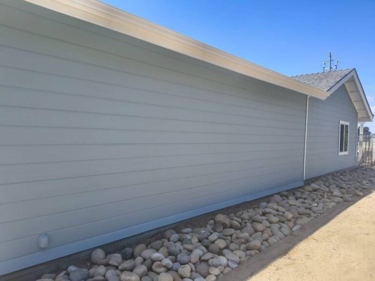 Side view of a house with newly painted light blue siding, white trim, and white gutters, featuring a decorative rock bed along the base under a clear blue sky. The house has a gray shingle roof and a small window.