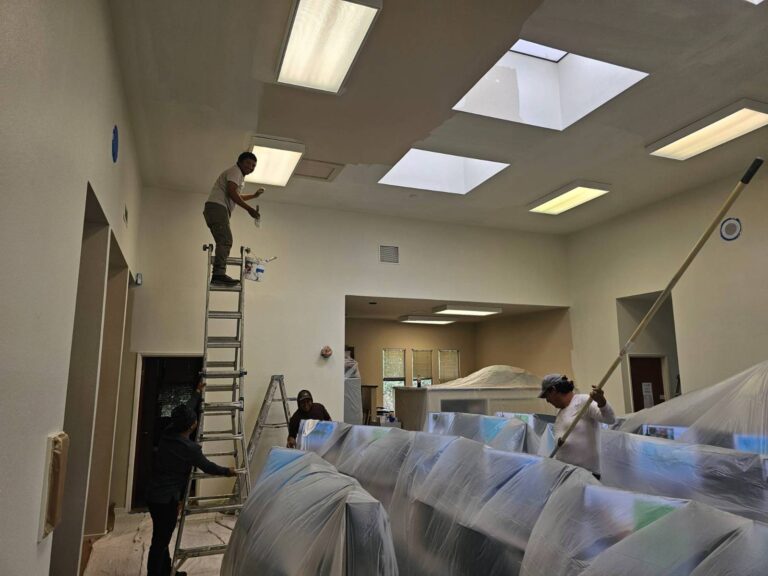 A team of professional painters from Hernandez Quality Paint working on a large interior ceiling. One painter stands on a tall ladder, carefully applying paint near a skylight, while another uses a long-handled roller to paint a lower section. The room's furniture is thoroughly protected with plastic sheeting, showcasing meticulous preparation for a commercial or residential painting project.