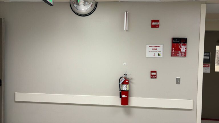 A light-colored wall in a hospital hallway featuring a red fire extinguisher, two red fire alarm pull stations, a 'NO PARKING ZONE GAS SHUT OFF/FIRE ALARM' sign, an emergency response guide, a convex security mirror, and a white handrail.