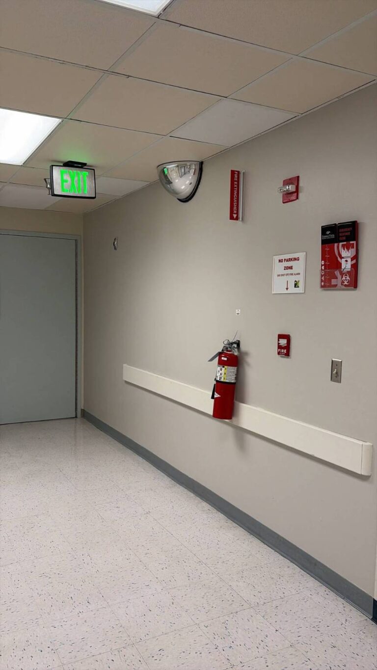 A well-lit hospital or office hallway with a green exit sign, a red fire extinguisher mounted on a white crash rail, a fire alarm, and a convex security mirror on the light gray wall. The floor is light speckled tile.