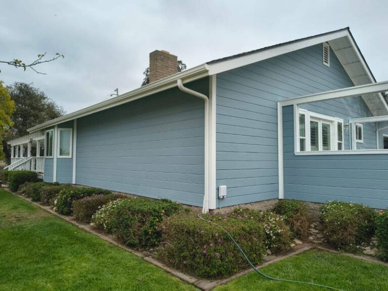 Exterior view of a house with newly painted light blue horizontal siding, white trim, and white gutters. A green lawn and landscaped bushes are in the foreground under an overcast sky.