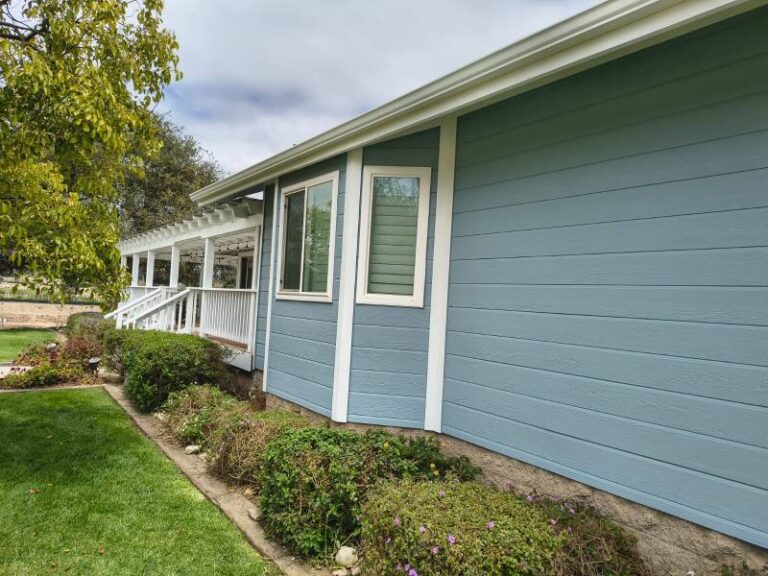 Exterior view of a house featuring light blue horizontal siding, crisp white window trim, and a white porch with railings. A green lawn and garden beds are visible in the foreground under an overcast sky.
