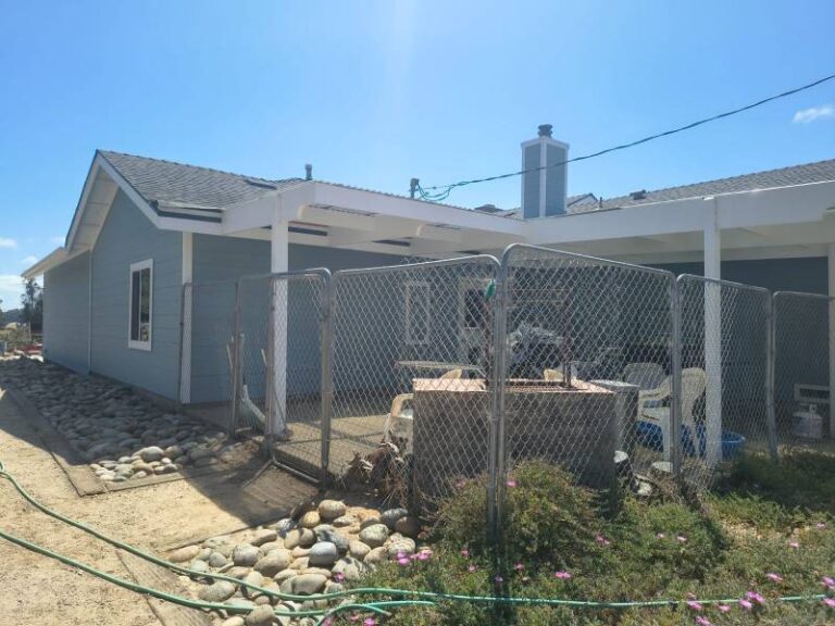 Exterior view of a house with new light blue siding, white trim, and a gray roof, featuring a patio area enclosed by a chain-link fence and rocky landscaping under a clear blue sky.