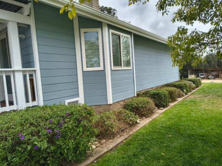 Side view of a house with light blue horizontal siding and white window trim, surrounded by green bushes and a well-maintained lawn under a cloudy sky.