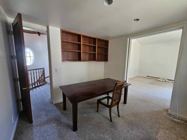An interior room featuring a dark wooden table with one chair, a built-in wooden shelf unit above the table, and light gray carpet. An open doorway on the left reveals a staircase and a round window, while another open doorway on the right leads to an empty room, both with matching carpet. The walls are light-colored, suggesting a space ready for painting or renovation.