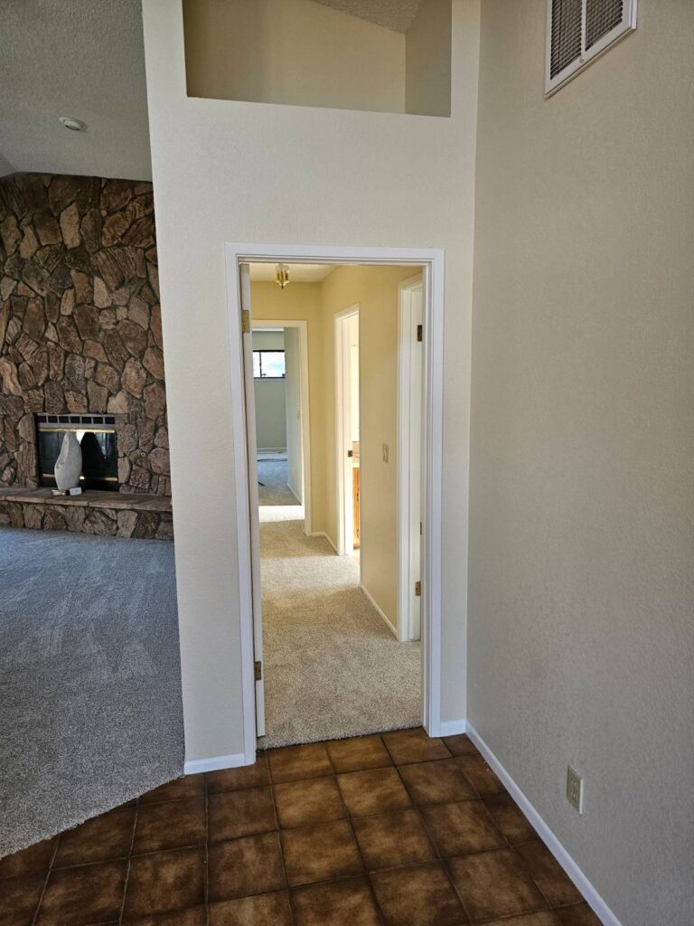 View from a tiled entryway looking into a carpeted hallway and a living room with a stone fireplace. The walls are painted a light, neutral color, showcasing a freshly painted interior.