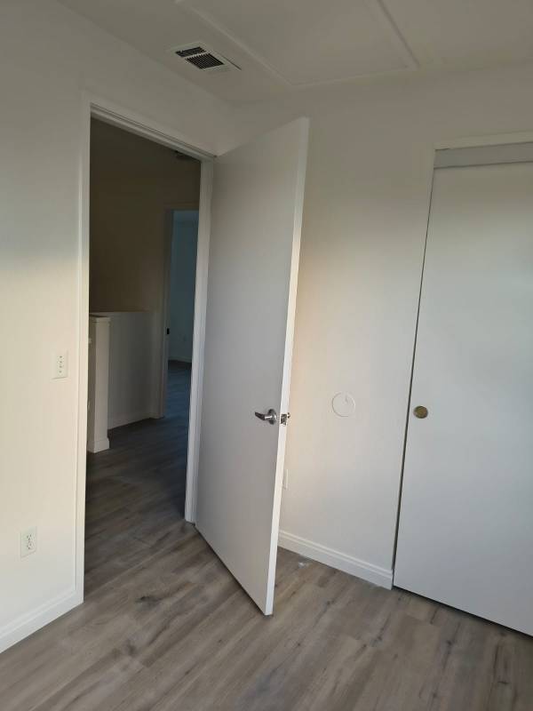 An interior room with freshly painted light gray walls, light wood-look laminate flooring, and a white door slightly ajar leading to a hallway. A white closet door is visible to the right.