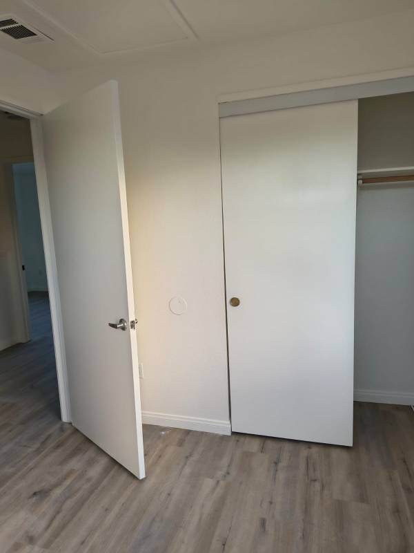 A clean, empty room featuring freshly painted white walls, light wood-look laminate flooring, a partially open white interior door with a silver handle, and a white sliding closet door with a gold knob revealing a closet rod.