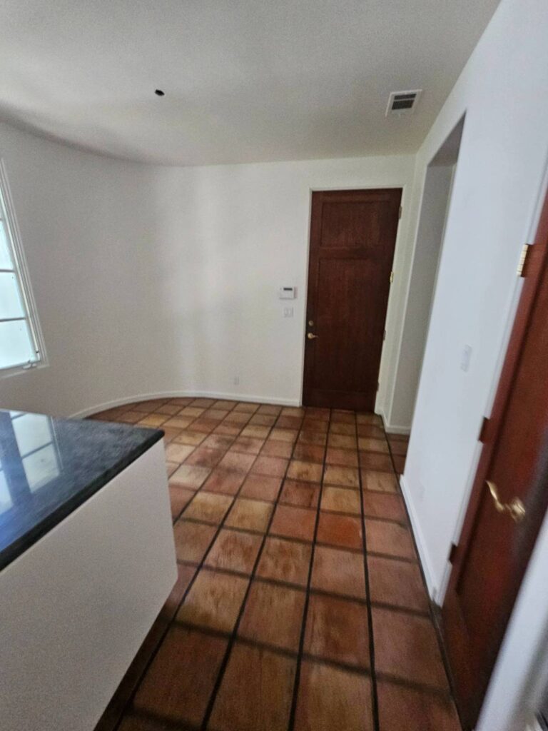 An empty interior room featuring freshly painted white walls, a dark brown wooden door, and a floor covered in rustic terracotta-style tiles. A dark countertop is visible on the left.