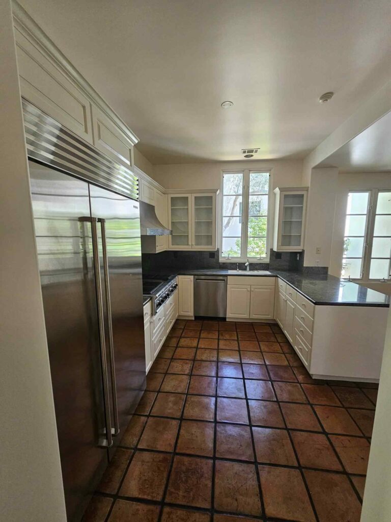 A spacious kitchen featuring white shaker-style cabinets, dark granite countertops, a large built-in stainless steel refrigerator, a gas range with a matching hood, and rustic terracotta floor tiles. Natural light fills the room through multiple windows.