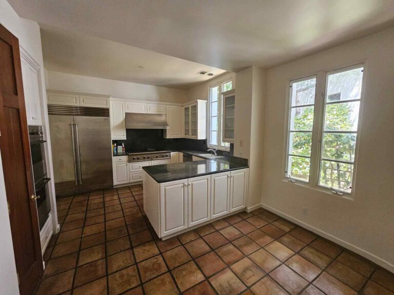 A spacious kitchen with white shaker-style cabinets, dark granite countertops, stainless steel refrigerator, double oven, and range hood, a kitchen island with a sink, and rustic terracotta-colored floor tiles laid diagonally. Large windows offer a view of outdoor greenery, and the walls are a light neutral color.