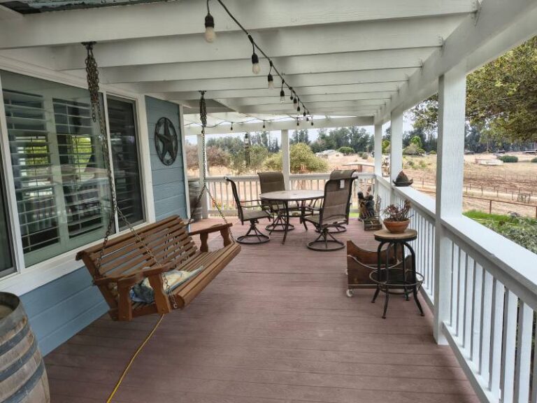 A spacious, freshly painted porch featuring light blue siding, white railings, and a white ceiling with string lights. A wooden porch swing hangs on the left, while an outdoor dining set with a round table and chairs is visible on the right. The porch overlooks a dry, rural landscape with fields and trees under a bright sky.